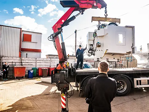 Crane unloading a large piece of three-phase equipment from a flatbed truck